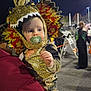 toddler, child, costume, dinosaur, pacifier, person, night, outdoor, holding, hoodie, face, headwear, event, people, parking_lot, lights, background, colorful, cute, fun