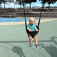 Ian joined the competition — help win amazing prizes! toddler, child, swing, playground, outdoor, sunny, trees, cityscape, blue_sky, shorts, tshirt, shoes, smiling, fun, daylight, shadow, leaves, rubber_floor, chain, bench
