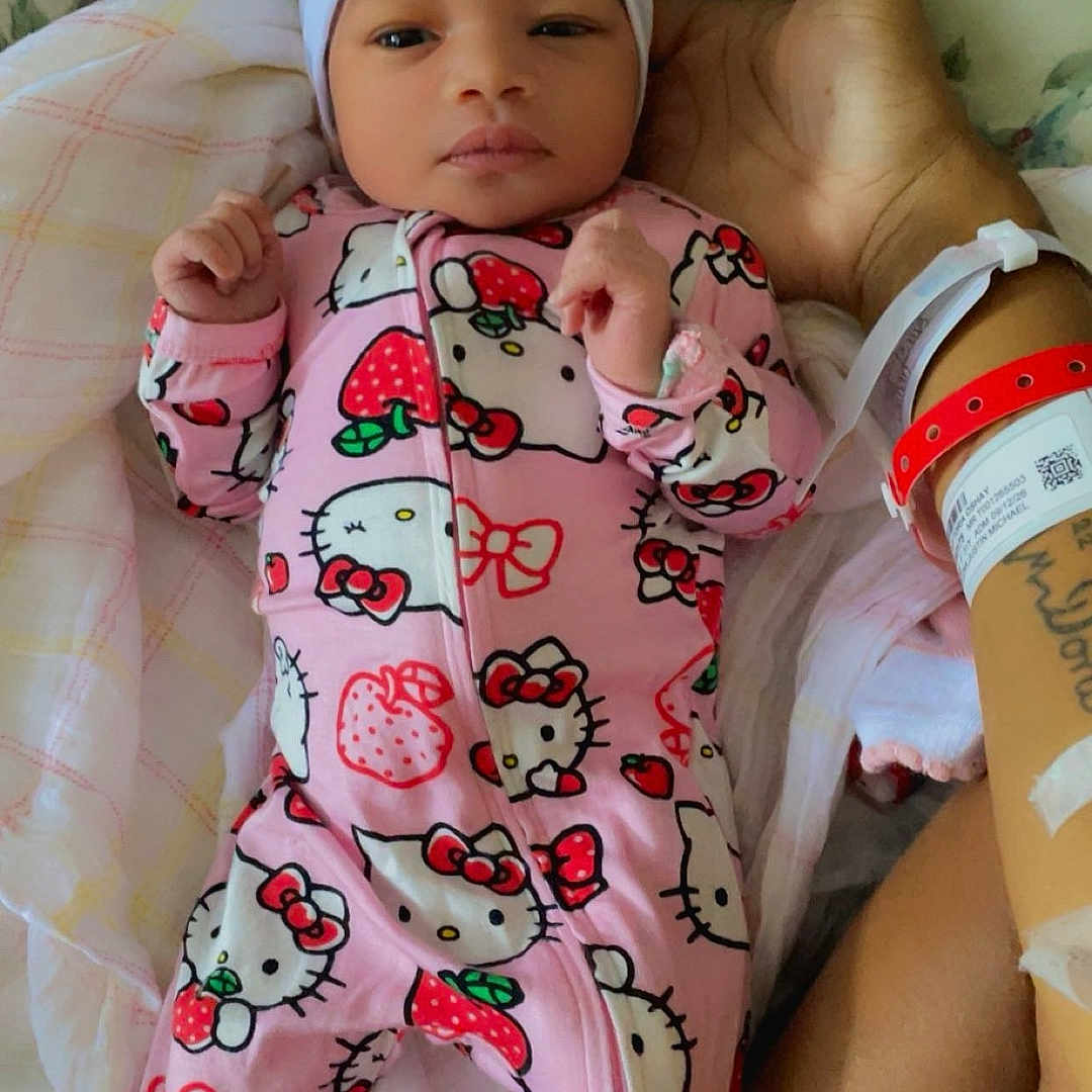 Stormi is registered to the contest to win money with this photo: baby, infant, child, portrait, lying_down, pink_onesie, hello_kitty, headband, bow, floral_bedding, blanket, hospital_bracelet, adult_arm, indoor, bed, cozy, serious_expression, hands, fashion, photography