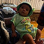baby, child, infant, person, clothing, hat, green_outfit, indoor, furniture, cabinet, hand, legs, feet, curious, seated, adult, holding, skull_pattern, fabric, home