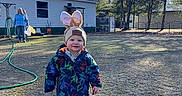 Walker is registered to the contest to win money with this photo: child, toddler, outdoor, grass, sunlight, jacket, hat, bunny_ears, smile, walking, person, adult, house, tree, hose, daylight, nature, playful, happy, winter_clothing