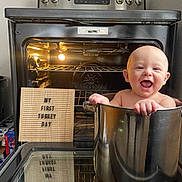 Walker is registered to the contest to win money with this photo: baby, smiling, pot, oven, kitchen, sign, reflection, happy, child, metallic_pot, cooking_pot, indoor, appliance, home, thanksgiving, holiday, light, window, playful