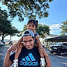 Sophia is registered to the contest to win money with this photo: child, man, smile, outdoor, daylight, trees, car, parking_lot, blue_sky, sandals, hat, tshirt, person, holding, shoulders, happy, family, playful, casual, people