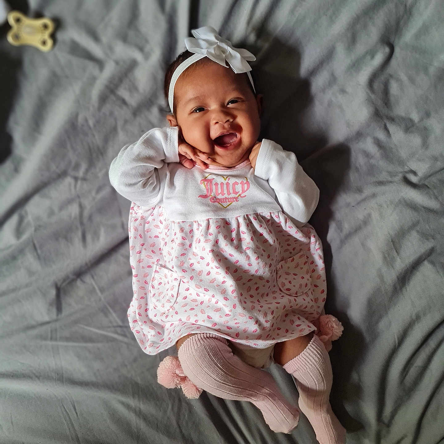 Lorelei-Rebecca joined the competition — help win amazing prizes! baby, bed, blanket, child, cozy, cute, dress, hands_near_face, happy, headband, indoors, infant, lying_down, pink_socks, playful, pom_poms, portrait, smiling, soft_lighting, white_clothing