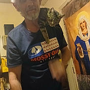 Ollie is registered to the contest to win money with this photo: animal, beard, blurred, cabinet, cap, casual, cat, door, furniture, home, indoor, man, person, pet, portrait, religious_art, shoulder, tabby_cat, tapestry, tshirt