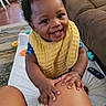 baby, carpet, chair, child, couch, fan, floor, hand, happy, indoor, legs, living_room, person, remote_control, skin, smiling, toy, window, wood_floor, yellow_bib