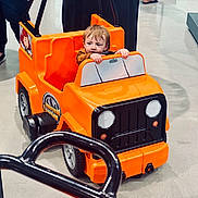 Phoebe is registered to the contest to win money with this photo: toddler, child, toy_car, orange, indoor, floor, person, hands, steering_wheel, plastic, vehicle, playing, curious, small, young_child, footwear, reflection, people, casual_clothing, shopping_area