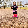 child, toddler, smiling, hoodie, boots, playground, wood_chips, outdoor, trees, blurred_background, person, recreation, fall, casual_clothing, happy, standing, daytime, clothing, footwear, nature