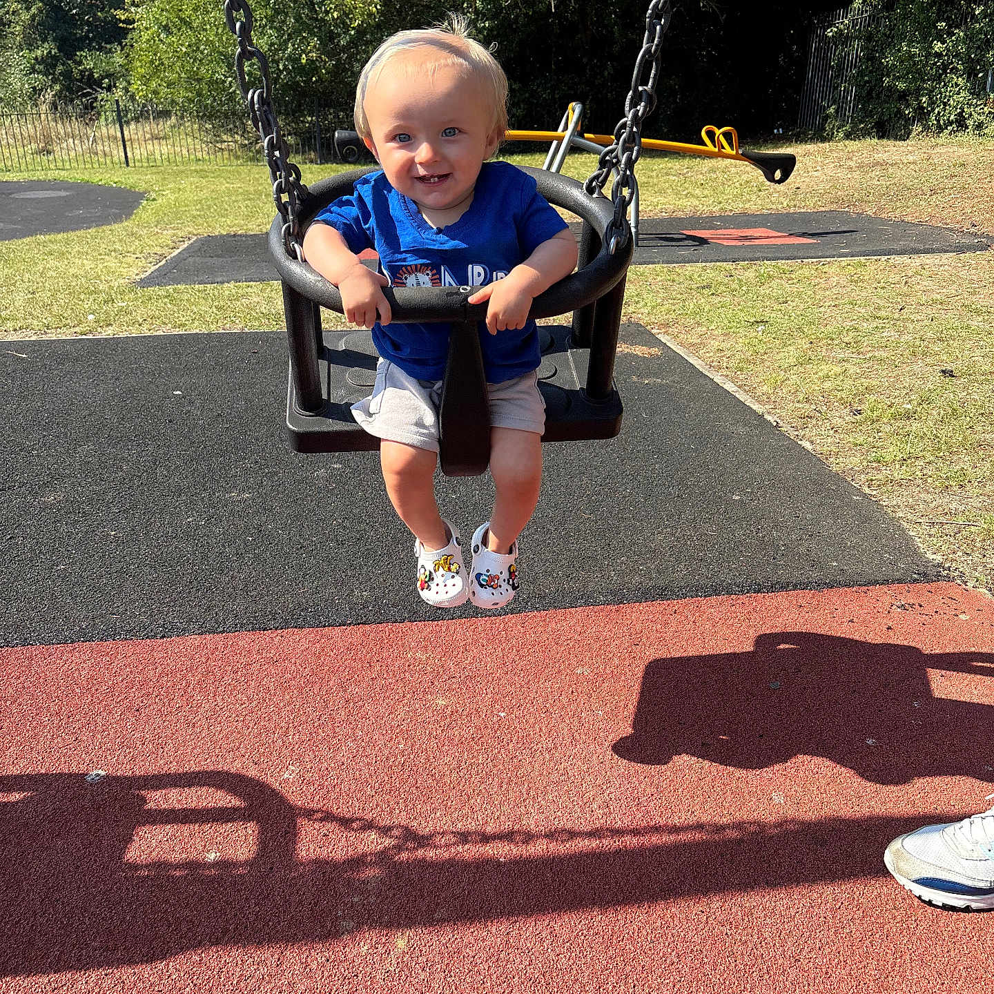 Reuben is registered to the contest to win money with this photo: blue_shirt, child, daylight, fun, grass, happy, metal_chain, outdoor, person, play, playground, shadow, shoes, shorts, smiling, summer, sunlight, swing, toddler, trees
