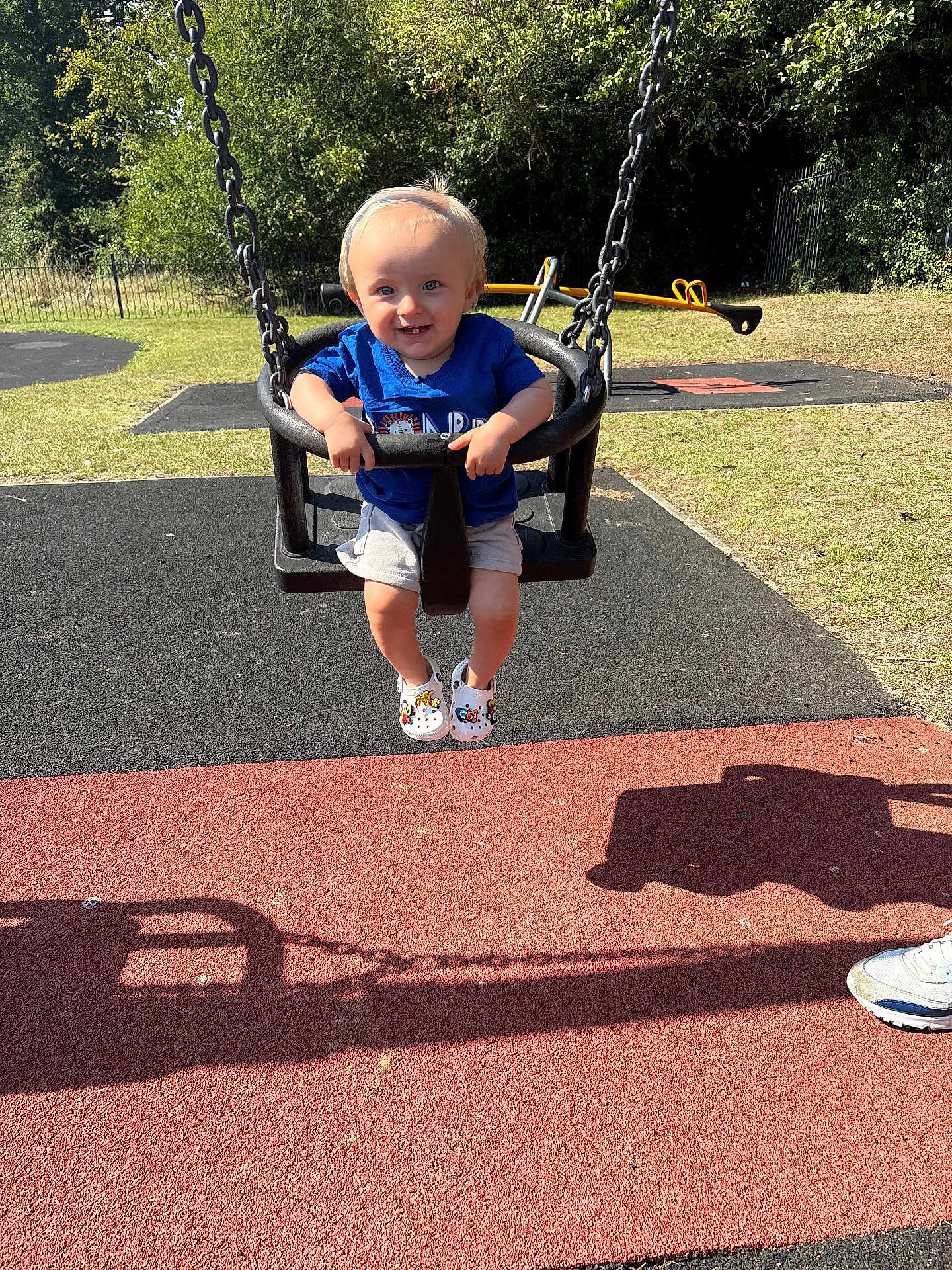 Reuben is registered to the contest to win money with this photo: toddler, child, swing, playground, smiling, blue_shirt, shorts, shoes, grass, trees, sunlight, shadow, outdoor, happy, person, play, summer, daylight, metal_chain, fun