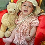 baby, teddy_bear, red_chair, pink_dress, lace, hat, pearl_necklace, bracelet, smile, happy, child, plush_toy, indoor, portrait, cute, sitting, furniture, accessories, person, decor