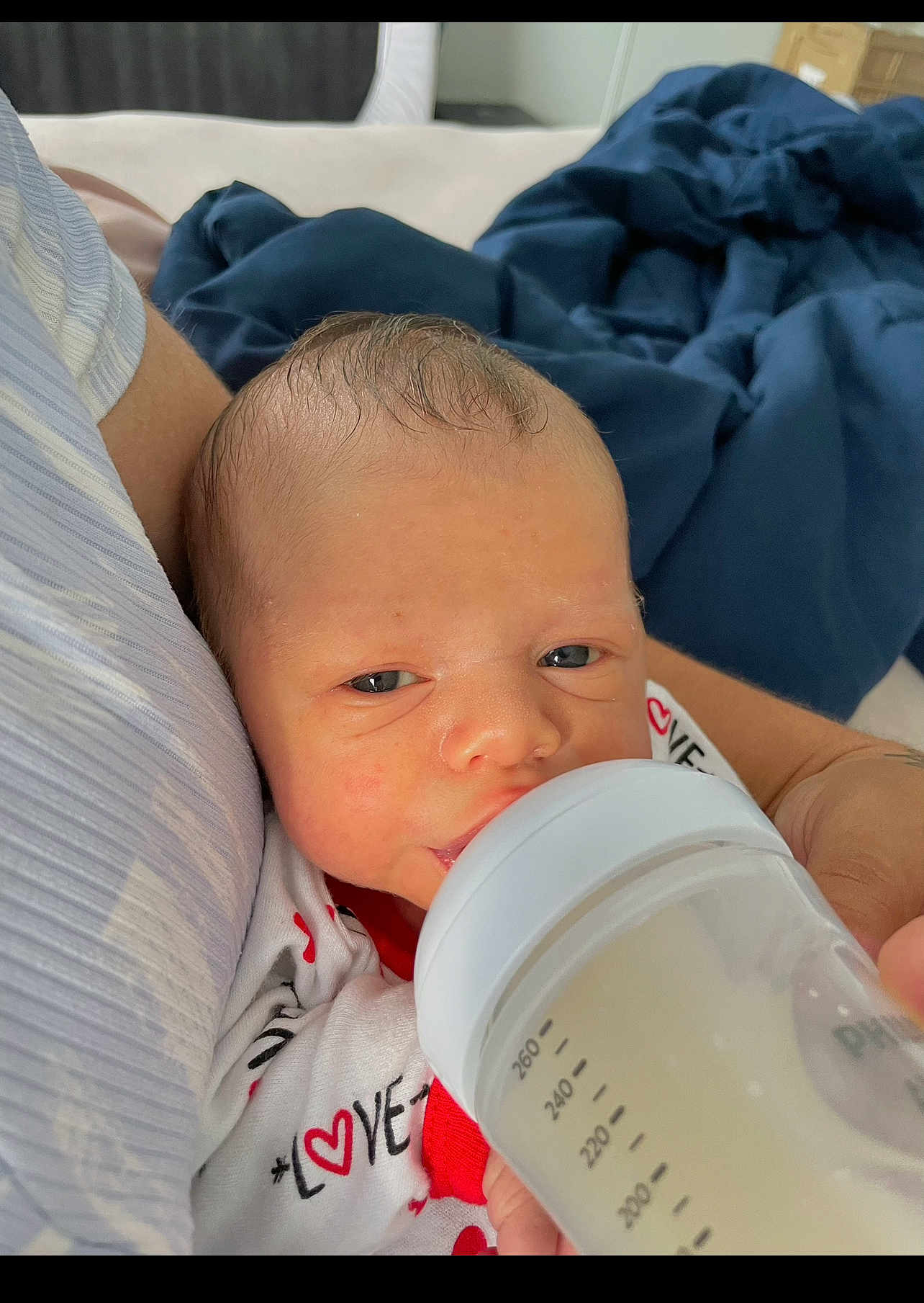 Luis joined the competition — help win amazing prizes! baby, newborn, bottle, feeding, infant, person, hand, clothing, blanket, arm, indoor, milk, onesie, love, face, head, sleepy, closeup, comfort, care