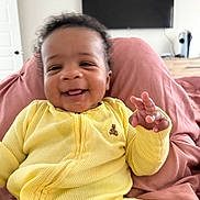 Aireon is registered to the contest to win money with this photo: baby, smiling, yellow_onesie, infant, blanket, indoor, cute, child, happy, person, home, furniture, portrait, sitting, cozy, soft, hand, face, young_child, relaxed