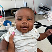 Airess is registered to the contest to win money with this photo: baby, smiling, infant, hand, living_room, white_onesie, happy, person, child, indoors, furniture, toy, floor, skin, face, expression, cute, small_child, holding, nail_polish