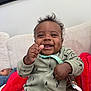 Airess is registered to the contest to win money with this photo: baby, smiling, onesie, pacifier_clip, red_blanket, tattooed_arm, couch, infant, happy, child, indoor, person, clothing, face, hand, sitting, furniture, blurred_background, two_children, home