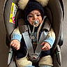 baby, car_seat, pacifier, knitted_clothing, hat, pom_poms, blue, cream, infant, child, sitting, cozy, warm, indoor, straps, safety_harness, cute, face, hands, footwear