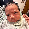 baby, infant, face, expression, furrowed_brow, onesie, hand, manicure, indoor, door, wall_decor, curly_hair, person, portrait, close_up, cozy, blanket, furniture, skin, clothing