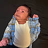 Benito is registered to the contest to win money with this photo: baby, infant, child, blue_plaid_shirt, white_onesie, clothing, lying_down, dark_background, portrait, cute, face, head, skin, hand, small_fist, newborn, person, indoors, casual, expression