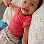 Rosalia is registered to the contest to win money with this photo: baby, bed, blanket, child, cozy, cute, dinosaur_print, expression, face, handwear, happy, indoors, infant, lying_down, person, pillow, pink_clothing, playful, smiling, young_child