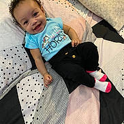 Rosalia is registered to the contest to win money with this photo: baby, smiling, curly_hair, blue_shirt, unicorn, bedspread, pillow, black_pants, socks, patterned_bed, happy, infant, child, indoors, cute, person, lying_down, bright_colors, cozy, portrait