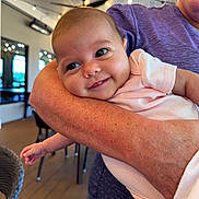 Madison is registered to the contest to win money with this photo: baby, smile, face, arm, person, indoor, lighting, string_lights, cheeky, happy, holding, clothing, pink_shirt, closeup, candid, infant, adult, warm_light, background, cozy