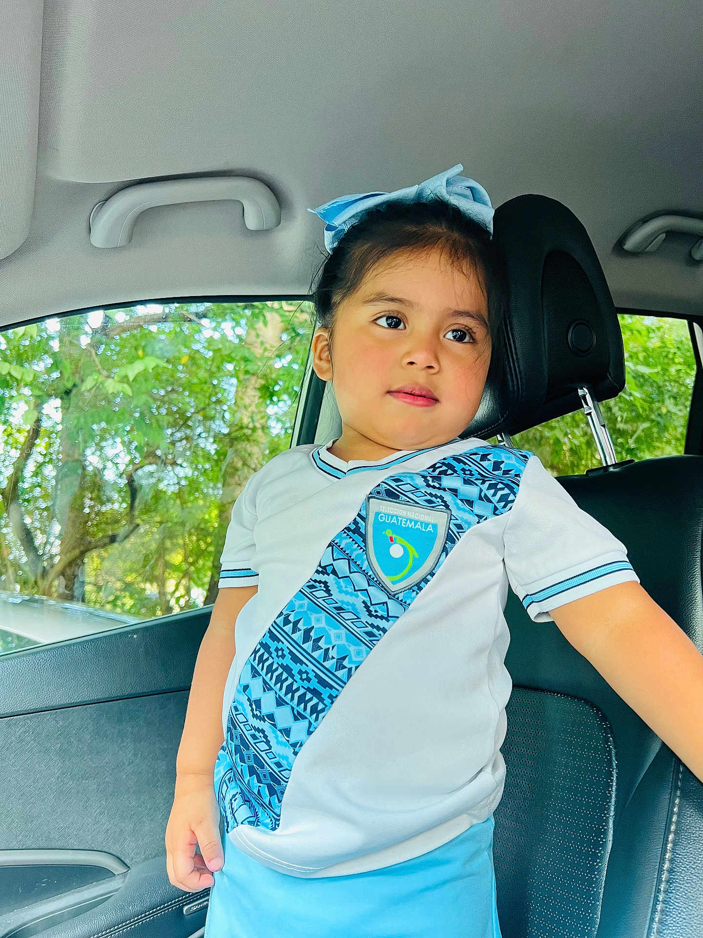 Arlet is registered to the contest to win money with this photo: blue_bow, car_interior, car_seat, casual, child, clothing, cute, daylight, expression, girl, guatemala, headwear, person, portrait, short_sleeves, soccer_jersey, standing, trees, window, young