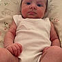 baby, infant, white_onesie, cushion, floral_pattern, indoor, person, skin, legs, arms, face, expression, sitting, natural_light, closeup, newborn, child, young, cute, comfort