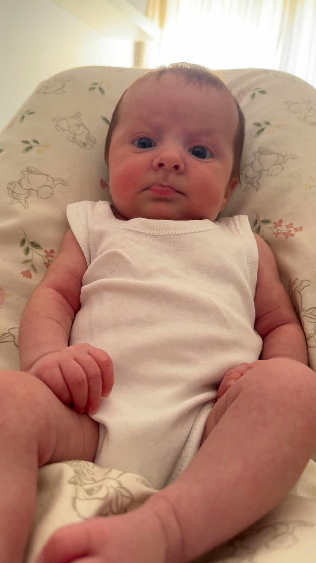 Arabella is registered to the contest to win money with this photo: baby, infant, white_onesie, cushion, floral_pattern, indoor, person, skin, legs, arms, face, expression, sitting, natural_light, closeup, newborn, child, young, cute, comfort