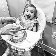 Sawyer is registered to the contest to win money with this photo: toddler, child, high_chair, cake, frosting, feeding, hands, smile, happy, indoor, footwear, shoes, floor, wall, power_outlet, decor, fabric, table, person, celebration