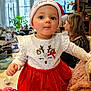baby, child, festive, santa_hat, red_dress, reindeer_design, indoor, woman, television, curious, holiday, cute, clothing, table, plant, bottle, toy, face, person, portrait