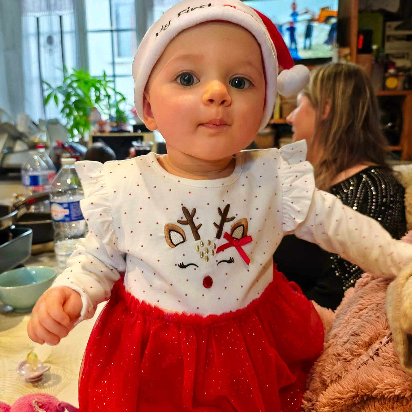 Shaïna a rejoint le concours — aidez-le/la à gagner de superbes lots ! baby, bottle, child, clothing, curious, cute, face, festive, holiday, indoor, person, plant, portrait, red_dress, reindeer_design, santa_hat, table, television, toy, woman