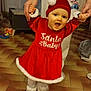 baby, child, red_dress, santa_hat, smiling, walking, indoor, floor, hands, helping, toy, home, person, footwear, white_socks, holiday, festive_clothing, happy, tiled_floor, family