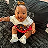 E Liah is registered to the contest to win money with this photo: baby, smiling, child, sitting, blanket, clothing, bow, happy, indoors, portrait, cute, footwear, toddler, black_blanket, graphic_tee, pink_shorts, bracelet, hair_tie, cozy, expression