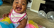 E Liah joined the competition — help win amazing prizes! baby, smiling, tutu, bib, colorful_clothing, sitting, blanket, balloons, toy, carpet, indoor, happy, face, child, person, living_room, curly_hair, home, cute, infant
