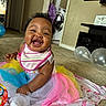 baby, smiling, tutu, bib, colorful_clothing, sitting, blanket, balloons, toy, carpet, indoor, happy, face, child, person, living_room, curly_hair, home, cute, infant