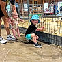 Jetsen joined the competition — help win amazing prizes! baby, child, man, petting_zoo, goat, fence, indoor, wooden_floor, barn, hat, shorts, sneakers, smile, animal, crouching, holding, people, livestock, daylight, casual_clothing