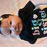baby, infant, headband, black_clothing, big_sister_text, smile, lying_down, cute, child, portrait, soft_surface, black_headband, onesie, young_child, face, baby_girl, colorful_text, happy, infant_fashion, close_up