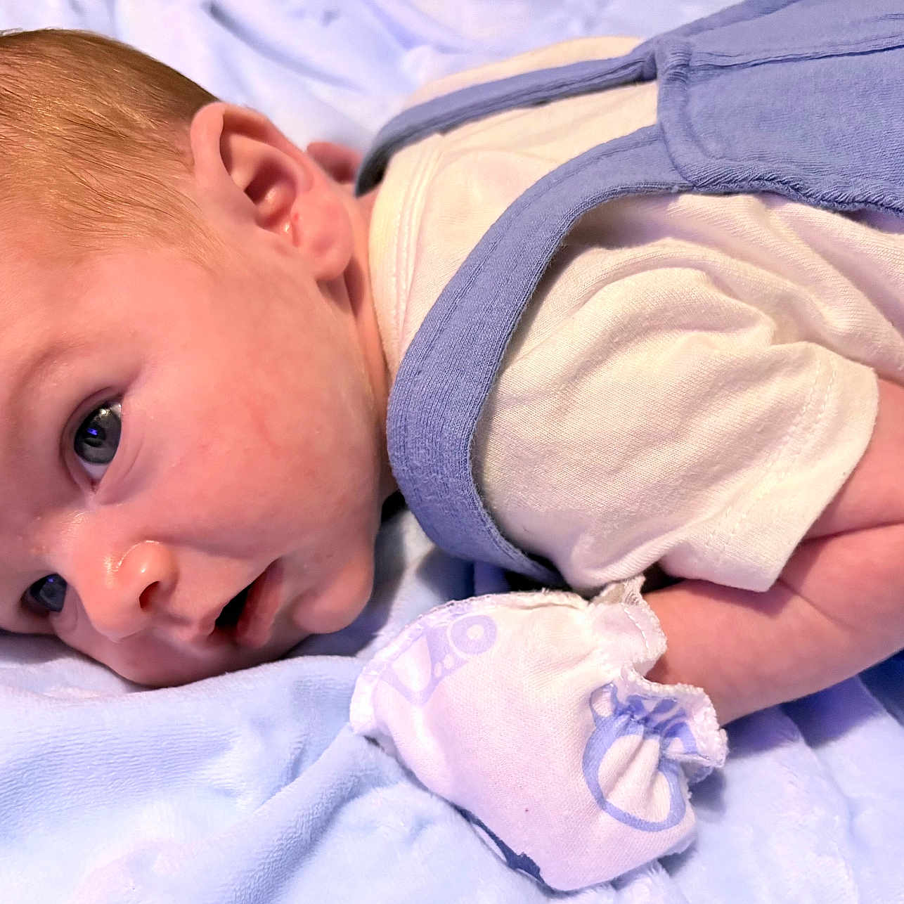 James is registered to the contest to win money with this photo: baby, infant, newborn, lying_down, blue_blanket, white_clothing, mitten, closeup, face, head, skin, ear, arm, cute, small, portrait, sleepwear, soft_texture, indoors, curious