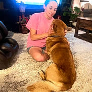 Charlie Booger is registered to the contest to win money with this photo: person, dog, carpet, living_room, furniture, television, indoor, petting, brown_dog, pink_shirt, casual_clothing, wooden_table, plant, sitting, relaxation, home, cozy, domestic_animal, interaction, comfort