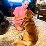 person, dog, carpet, living_room, furniture, television, indoor, petting, brown_dog, pink_shirt, casual_clothing, wooden_table, plant, sitting, relaxation, home, cozy, domestic_animal, interaction, comfort