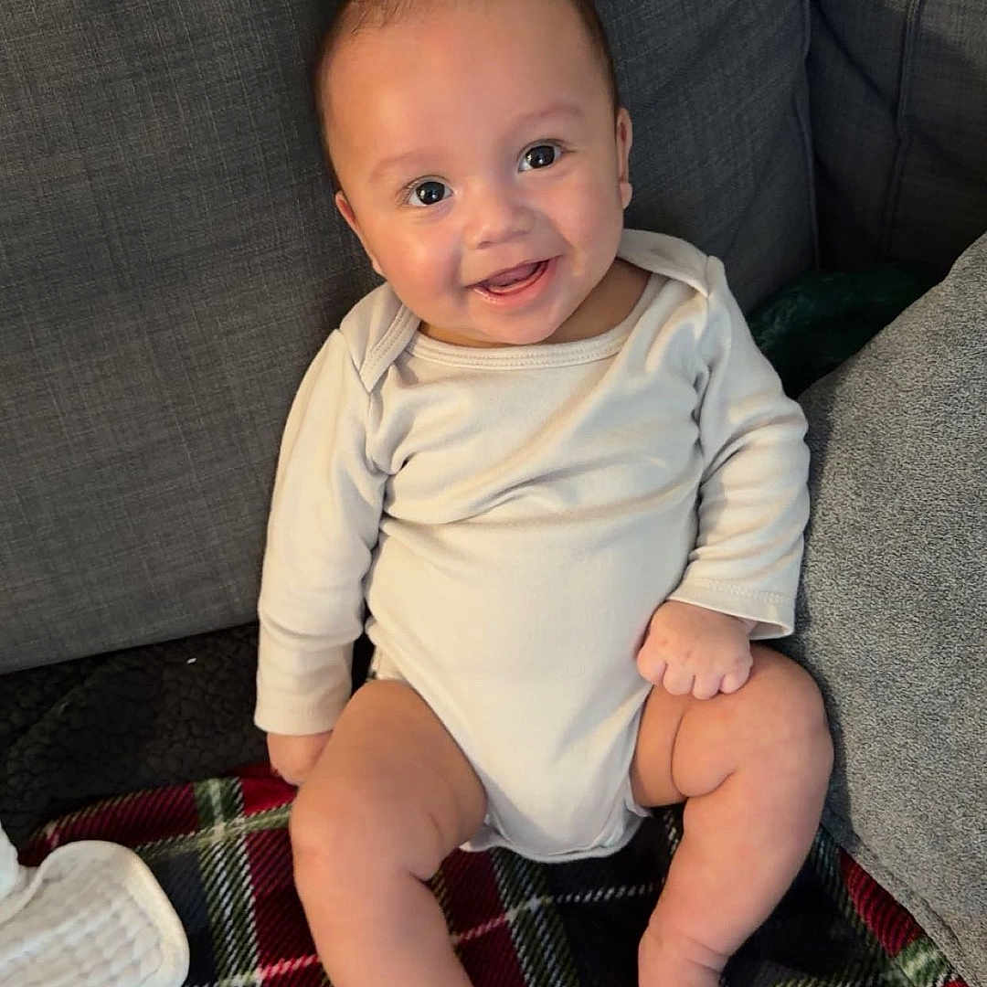 Lucas is registered to the contest to win money with this photo: baby, smiling, infant, sitting, blanket, plaid, couch, gray, happy, cute, child, indoors, face, skin, foot, hand, onesie, cozy, comfort, young