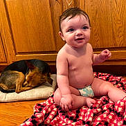 Madison joined the competition — help win amazing prizes! baby, child, infant, sitting, smiling, face, portrait, dog, sleeping_dog, pet, blanket, red_blanket, cushion, wooden_cabinets, indoor, home, floor, barefoot, diaper, cozy