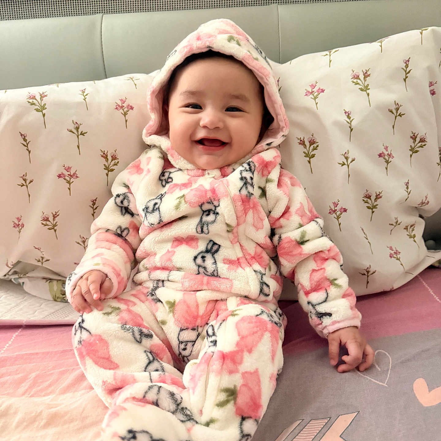 Keira is registered to the contest to win money with this photo: baby, child, smiling, happy, portrait, indoor, bed, pillow, bedding, floral, pink, hood, onesie, fleece, rabbit_pattern, sitting, infant, face, cute, bedroom