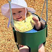 Raelynn is registered to the contest to win money with this photo: baby, child, swing, playground, hat, bib, sunlight, smile, happy, outdoor, woodchips, green, seat, metal, summer, cute, infant, fun, play, park