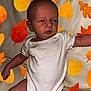 Arlo is registered to the contest to win money with this photo: baby, infant, white_onesie, autumn_leaves, fall, background_pattern, skin, arm, leg, face, expression, indoor, couch, seated, child, young, cute, person, pattern, colorful