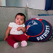 Gabriella is registered to the contest to win money with this photo: air_conditioner, baby, blue_curtain, child, couch, cushion, dark_hair, football, home, indoors, infant, patriots, pillow, pink_socks, portrait, red_pants, sitting, sofa, sports, wide_eyes