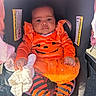 baby, car_seat, child, costume, cute, daylight, eyes, face, halloween, indoor, infant, orange_clothing, photography, portrait, pumpkin_costume, safety_harness, sitting, striped_pants, vehicle, white_blanket