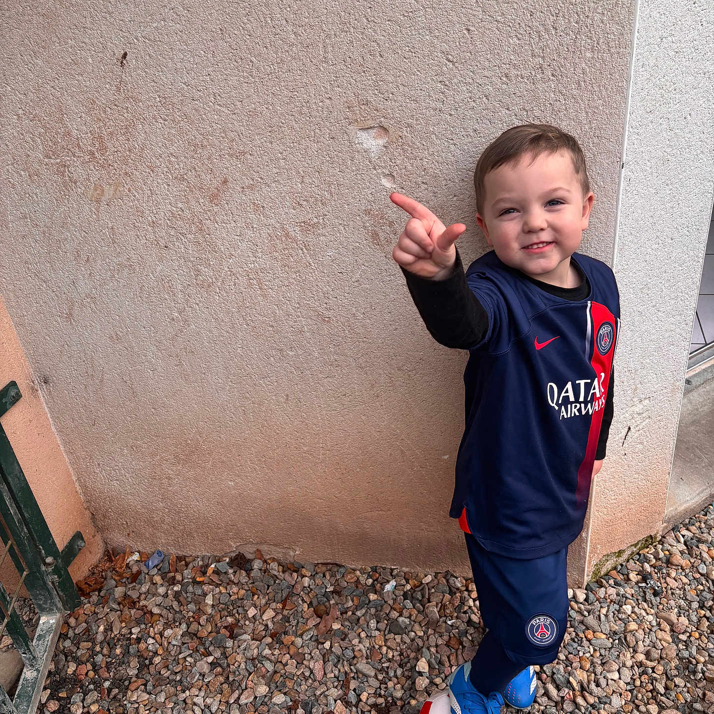Loïs participe au concours pour gagner de l'argent avec cette photo : ball, blue_clothing, boy, casual, child, football, footwear, gate, gravel, outdoor, playful, pointing, portrait, psg, red, smiling, soccer, sportswear, wall, white