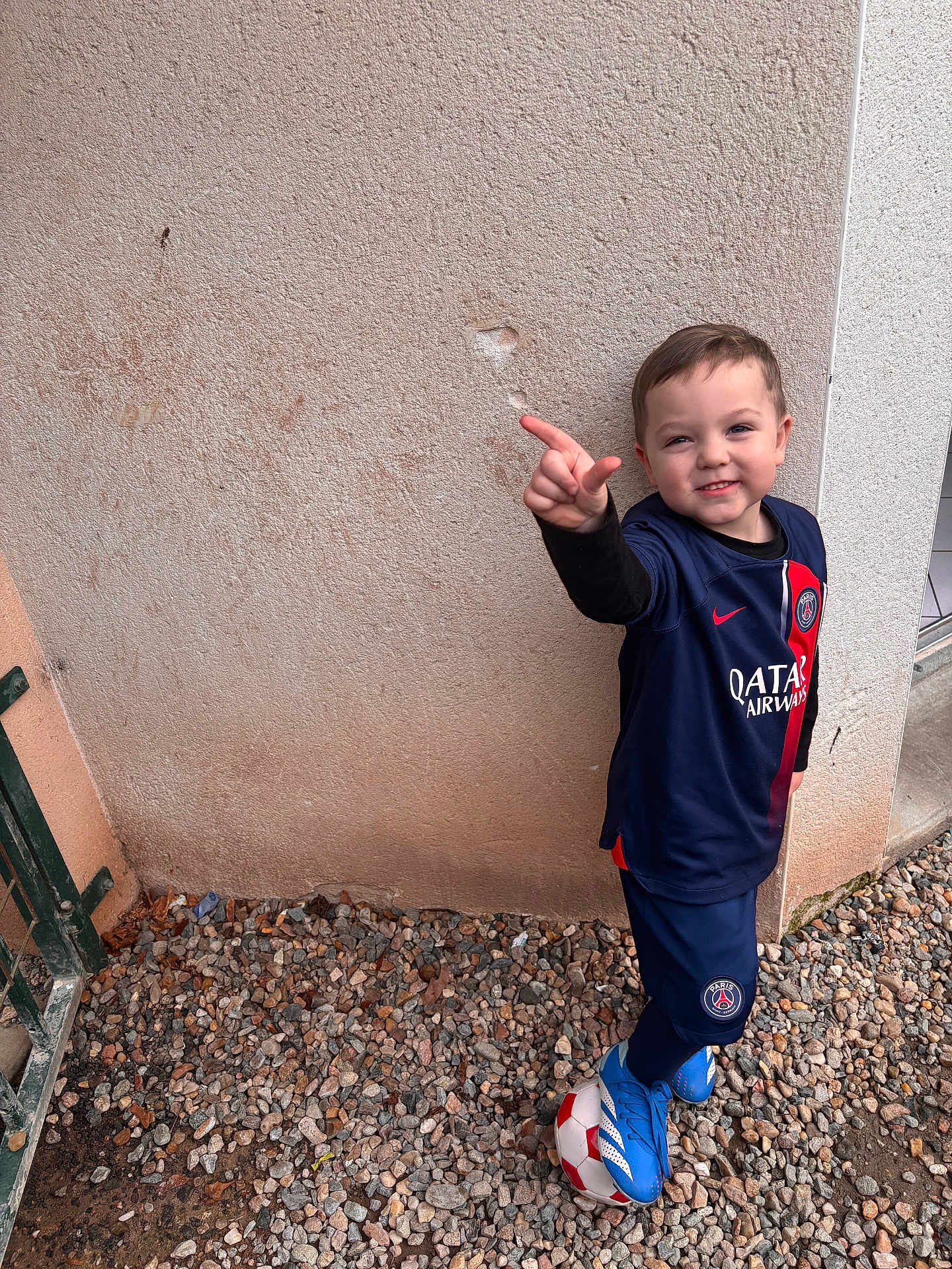 Loïs participe au concours pour gagner de l'argent avec cette photo : ball, blue_clothing, boy, casual, child, football, footwear, gate, gravel, outdoor, playful, pointing, portrait, psg, red, smiling, soccer, sportswear, wall, white