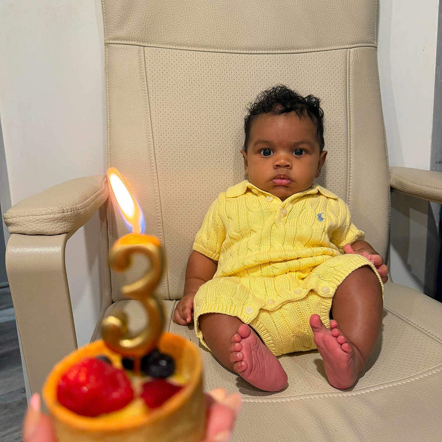 Genuine is registered to the contest to win money with this photo: baby, child, birthday, candle, cake, yellow_clothing, chair, leather_chair, celebration, indoor, portrait, serious_expression, sitting, hand, dessert, fruit, strawberry, blueberry, knitwear, cute
